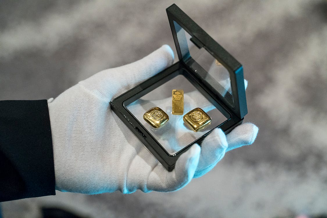 a person wearing gloves holding a glass frame with gold bars - How to Choose the Right Gold Assets for Your Goals